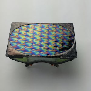 Op de afbeelding: Een kleine, rechthoekige glazen doos met een iriserend, ovaalvormig deksel. Het deksel heeft een golvend patroon en is ingelijst met zwart. De doos heeft een groene basis en staat op vier kleine, koperkleurige voetjes.