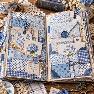May include: An open scrapbook featuring blue and white patchwork fabric pages with pockets. It contains floral-patterned cards, tags with blue gingham ribbons, lace trim, and blue wax seals. One card features the text Collect Beautiful Moments.