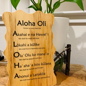 May include: A wooden sign with a Hawaiian proverb, "Aloha Oli", written in black lettering. The proverb is written in English and Hawaiian, with the English translation below each line. The sign is on a wooden surface.