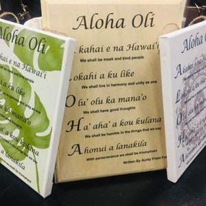 May include: Three wooden signs with the Hawaiian phrase "Aloha Oli" and a traditional Hawaiian chant written in English below. The signs are painted in different colors and feature a variety of designs, including a green monstera leaf, a gray floral pattern, and a white background with a floral design.