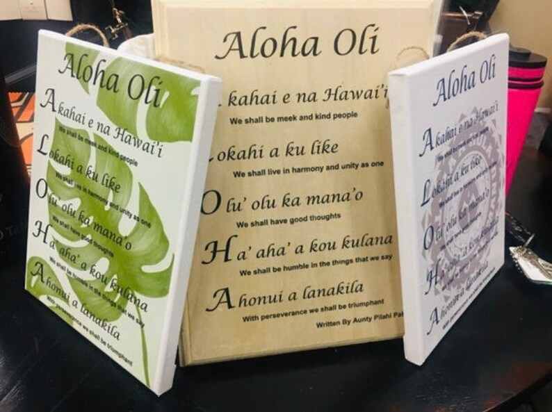 May include: Three wooden signs with the Hawaiian phrase "Aloha Oli" and a traditional Hawaiian chant written in English below. The signs are painted in different colors and feature a variety of designs, including a green monstera leaf, a gray floral pattern, and a white background with a floral design.
