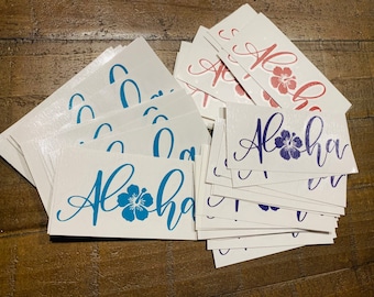 May include: A stack of white stickers with the word "Aloha" written in blue, purple, and red cursive lettering. Each sticker features a stylized hibiscus flower in the corresponding color.
