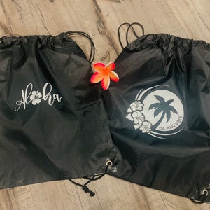 May include: Two black drawstring bags with white designs. One bag says "Aloha" with a white hibiscus flower. The other bag has a white circle with a palm tree and two hibiscus flowers and the text "Island Vibes".