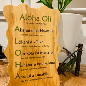 May include: Wooden sign with a Hawaiian theme, featuring the phrase "Aloha Oli" and a list of Hawaiian values, including "Akahai e na Hawai'i", "Lōkahi a kūlike", "Olu Olu ka mana o", "Ha`aha`a kou kūlana", and "Ahonui a Lanakila".