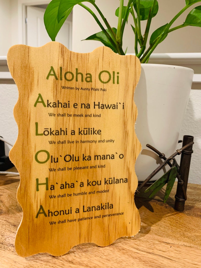 May include: Wooden sign with a Hawaiian theme, featuring the phrase "Aloha Oli" and a list of Hawaiian values, including "Akahai e na Hawai'i", "Lōkahi a kūlike", "Olu Olu ka mana o", "Ha`aha`a kou kūlana", and "Ahonui a Lanakila".