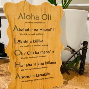 May include: Wooden sign with the Hawaiian phrase "Aloha Oli" and the text "Written by Aunty Pilahi Paki". The sign lists five Hawaiian words and their meanings in English: Akahai e na Hawai'i (We shall be meek and kind), Lōkahi a kūlike (We shall live in harmony and unity), Olu Olu ka mana o (We shall be pleasant and kind), Ha`aha`a kou kūlana (We shall be humble and modest), and Ahonui a Lanakila (We shall have patience and perseverance).