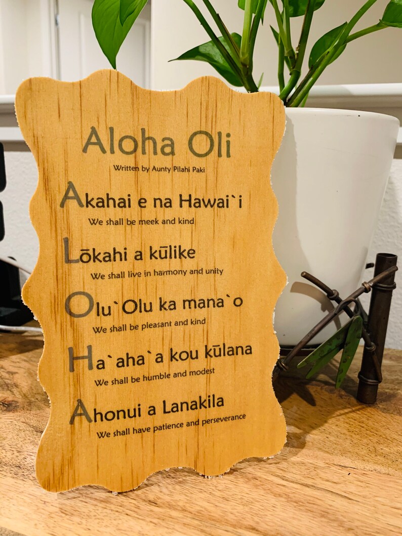 May include: Wooden sign with the Hawaiian phrase "Aloha Oli" and the text "Written by Aunty Pilahi Paki". The sign lists five Hawaiian words and their meanings in English: Akahai e na Hawai'i (We shall be meek and kind), Lōkahi a kūlike (We shall live in harmony and unity), Olu Olu ka mana o (We shall be pleasant and kind), Ha`aha`a kou kūlana (We shall be humble and modest), and Ahonui a Lanakila (We shall have patience and perseverance).