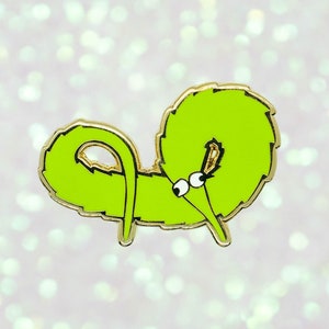 May include: A lime green enamel pin depicting a cartoon worm with a gold outline. The worm has a curly body, googly eyes, and a small gold accent. The pin is set against a sparkly, light background.