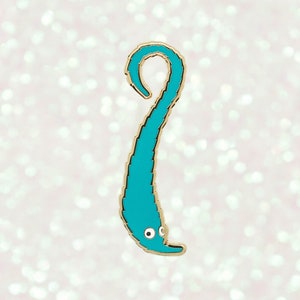 May include: A teal worm pin with a gold outline and small white eyes. The worm is in a curved shape, with a textured body. The pin is set against a soft, glittery background.