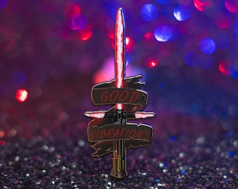 May include: A metal pin featuring a red lightsaber design with the words "GOOD VIBRATIONS" on a signpost. The pin has a gold base and is set against a glittery background with red and blue bokeh.
