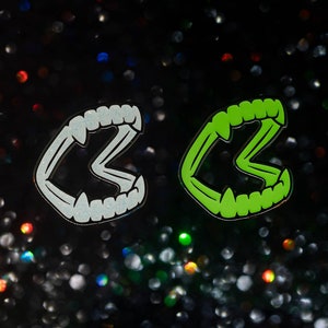 May include: Two enamel pins in the shape of stylized vampire fangs. One pin is white with glitter, the other is bright green. Both have black outlines and are set against a dark, glittery background.