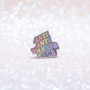 May include: A silver-colored enamel pin with the words "ANTI ANTI VAXXER CLUB" in rainbow colors. The pin is set against a sparkly, glittery background.