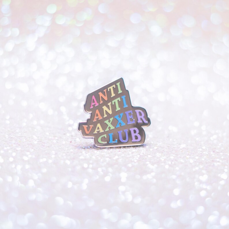May include: A silver-colored enamel pin with the words "ANTI ANTI VAXXER CLUB" in rainbow colors. The pin is set against a sparkly, glittery background.