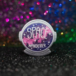 Peut inclure: Écusson brodé avec les mots "SPACE WANDERER" en lettres blanches. L'écusson présente une scène spatiale violette et rose avec des étoiles et une bordure blanche. L'écusson est rond avec un fond incurvé.