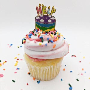 K&ouml;nnte beinhalten: Ein Cupcake mit rosa Zuckerguss und bunten Streuseln. Eine kuchenf&ouml;rmige Anstecknadel mit dem Wort "HELP" oben drauf. Die Anstecknadel hat ein Regenbogen-Design und goldfarbene Akzente. Der Cupcake befindet sich auf wei&szlig;em Hintergrund.