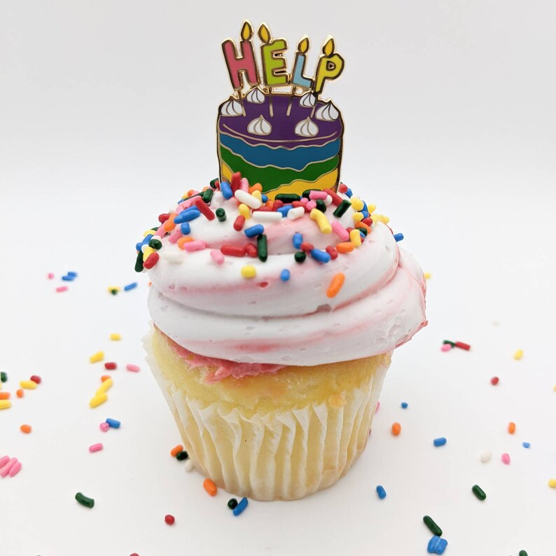 K&ouml;nnte beinhalten: Ein Cupcake mit rosa Zuckerguss und bunten Streuseln. Eine kuchenf&ouml;rmige Anstecknadel mit dem Wort "HELP" oben drauf. Die Anstecknadel hat ein Regenbogen-Design und goldfarbene Akzente. Der Cupcake befindet sich auf wei&szlig;em Hintergrund.
