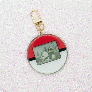 May include: A round keychain with a gold clasp. The keychain features a red and white design with a gray rectangular image of a cartoon scene. The scene includes a sun, trees, and a cartoon character. The number "09" is visible.
