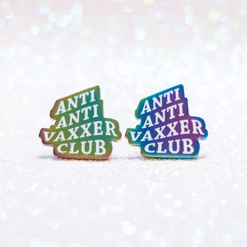 May include: Two iridescent enamel pins with the words "ANTI ANTI VAXXER CLUB" in a stacked design. The pins have a gradient of colors, including green, blue, and purple. The background is a sparkly, glittery white.
