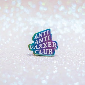 May include: A colorful pin with the words "ANTI ANTI VAXXER CLUB" in white lettering. The pin has a gradient of colors, including blue, green, and purple. The background is a sparkly, glittery surface.