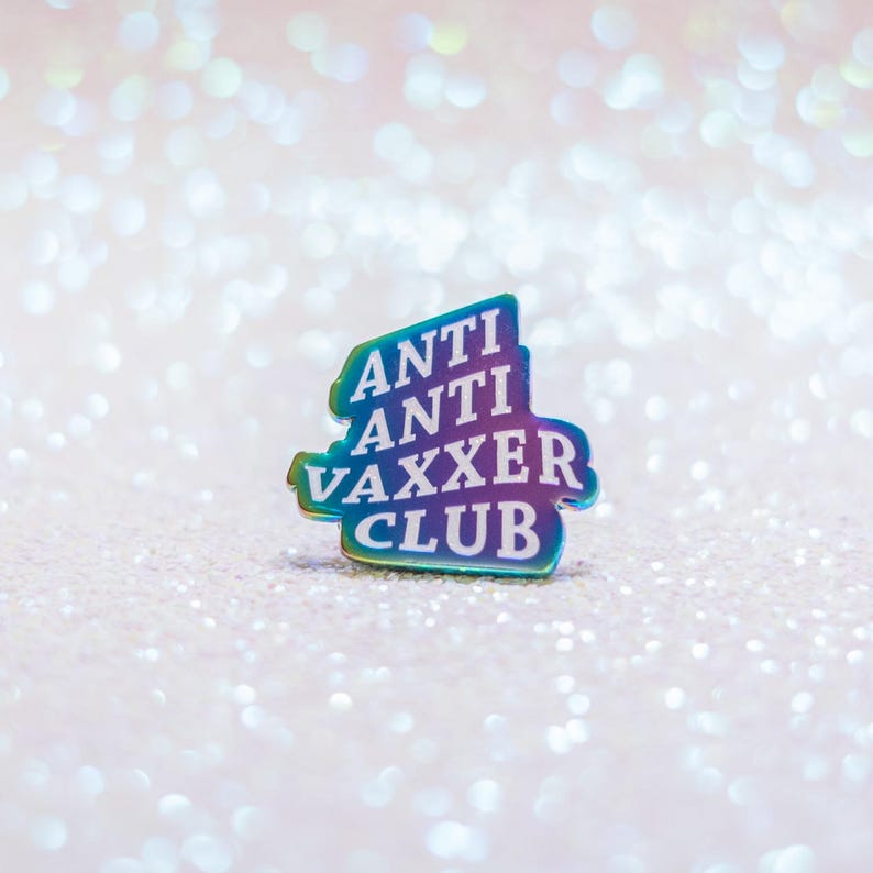 May include: A colorful pin with the words "ANTI ANTI VAXXER CLUB" in white lettering. The pin has a gradient of colors, including blue, green, and purple. The background is a sparkly, glittery surface.