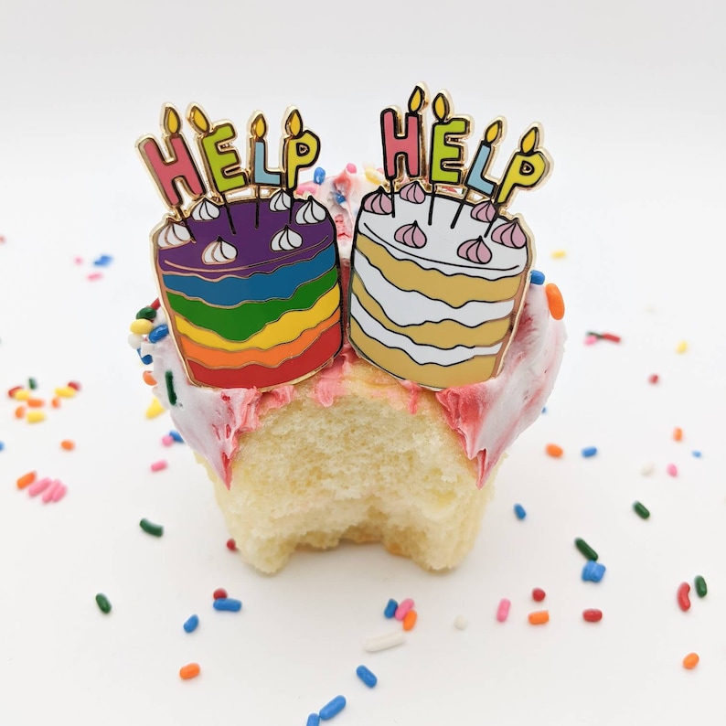 K&ouml;nnte beinhalten: Zwei Emaille-Anstecknadeln in Kuchenform, jede mit dem Wort "HELP" oben. Einer ist regenbogenfarben, der andere wei&szlig; mit rosa Akzenten. Die Anstecknadeln befinden sich auf einem Cupcake mit rosa Zuckerguss und Streuseln.