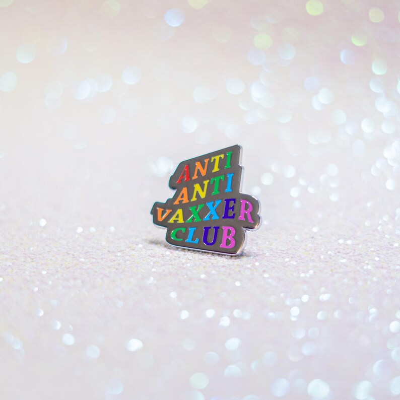 May include: A silver-colored enamel pin with the words "ANTI ANTI VAXXER CLUB" in rainbow colors. The pin is in a stylized, layered design, set against a sparkly, light-colored background.