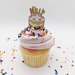 K&ouml;nnte beinhalten: Ein Cupcake mit wei&szlig;em Zuckerguss und bunten Streuseln. Eine kuchenf&ouml;rmige Anstecknadel mit dem Wort "HELP" darauf. Der Cupcake hat eine gelbe Basis und befindet sich auf wei&szlig;em Hintergrund.