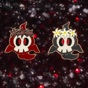 Op de afbeelding: Twee emaille pins met een spookachtig schedelontwerp. De ene pin is rood met een bruine bloemenkrans, de andere is zwart met een witte en gele bloemenkrans. Beide hebben goudkleurige contouren en rode ogen.