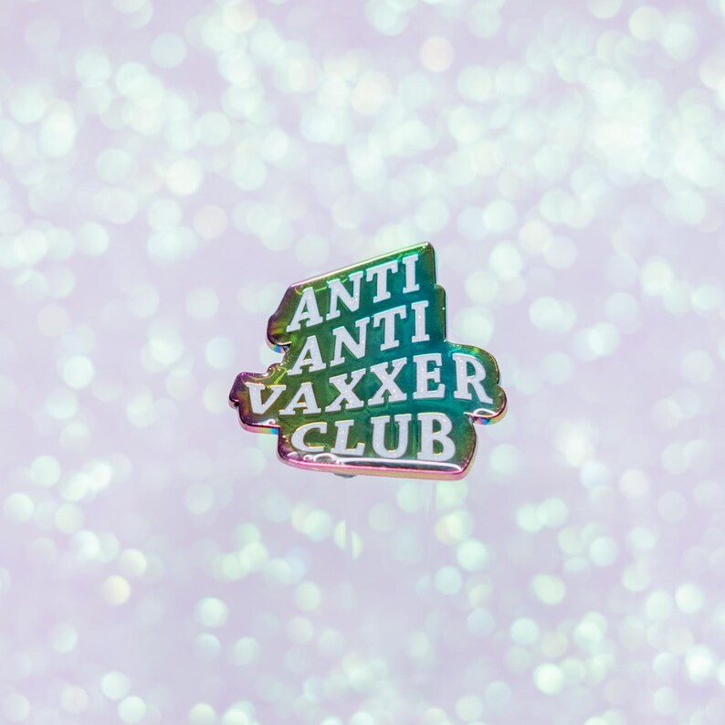 May include: A colorful enamel pin with the words "ANTI ANTI VAXXER CLUB" in white lettering. The pin has a gradient of green, blue, and gold colors. The background is a soft, out-of-focus, light purple with white bokeh.