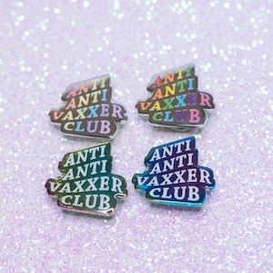 May include: Four enamel pins with the text "ANTI VAXXER CLUB" in a rainbow gradient color scheme. The pins are on a sparkly, light purple surface.
