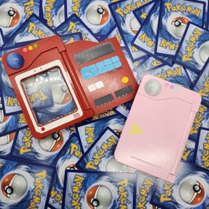 Op de afbeelding: Drie kleurrijke, draagbare elektronische apparaten, rood, roze en rood, met knoppen en schermen, worden weergegeven op een achtergrond van Pokémon-verzamelkaarten. Het rode apparaat is open en onthult een helder venster. Het roze apparaat is gesloten.