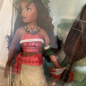 May include: A Moana doll with long black hair, wearing a brown and white skirt, a red sash, and a green lei. She is holding a brown wooden staff with red feathers.