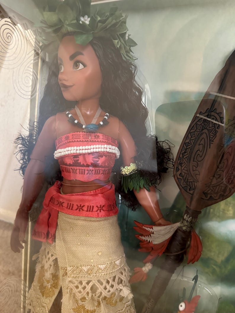 May include: A Moana doll with long black hair, wearing a brown and white skirt, a red sash, and a green lei. She is holding a brown wooden staff with red feathers.