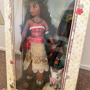 May include: A limited edition Moana doll with a flower crown, red and white top, and a brown skirt. The doll is holding a wooden paddle and is standing next to a small pig and a rooster. The box is decorated with a floral pattern and the text "Disney Moana Limited Edition Limited to 6500".
