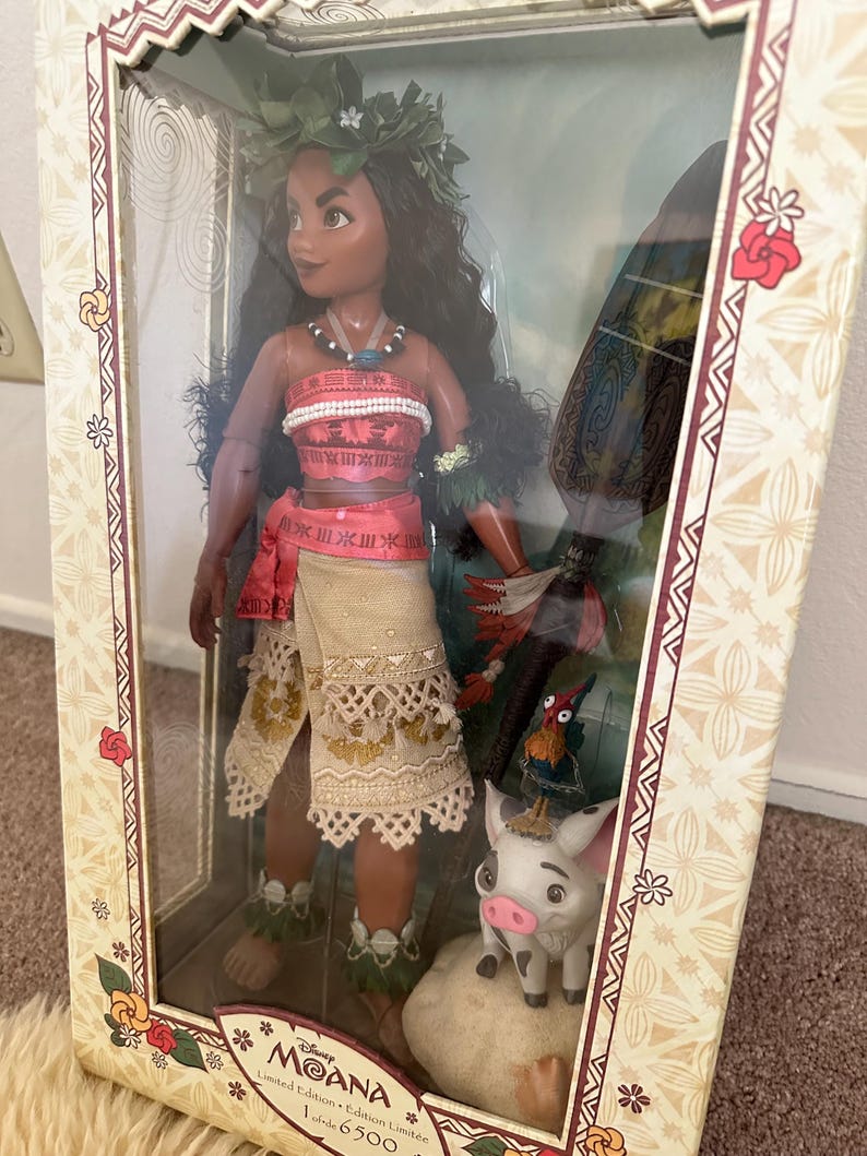 May include: A limited edition Moana doll with a flower crown, red and white top, and a brown skirt. The doll is holding a wooden paddle and is standing next to a small pig and a rooster. The box is decorated with a floral pattern and the text "Disney Moana Limited Edition Limited to 6500".