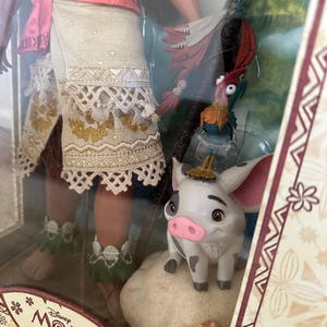May include: A limited edition Moana doll, part of a collection of 6,500. The doll is wearing a brown skirt with a white lace trim and a pink top. She is holding a brown staff with a green leaf on top. The doll is standing next to a brown pig with a pink nose and a white and brown rooster with red feathers.
