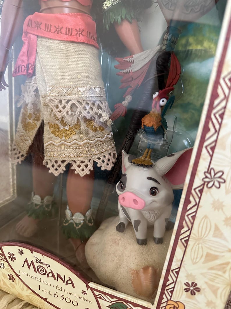 May include: A limited edition Moana doll, part of a collection of 6,500. The doll is wearing a brown skirt with a white lace trim and a pink top. She is holding a brown staff with a green leaf on top. The doll is standing next to a brown pig with a pink nose and a white and brown rooster with red feathers.