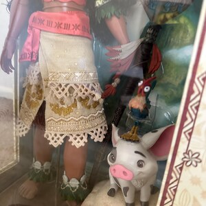 May include: A Disney Moana doll with brown skin, wearing a red and white top, a brown skirt with white trim, and a green lei. She is holding a spear and standing next to a white pig with black spots. The pig is standing on a rock. The background is a brown and white pattern.