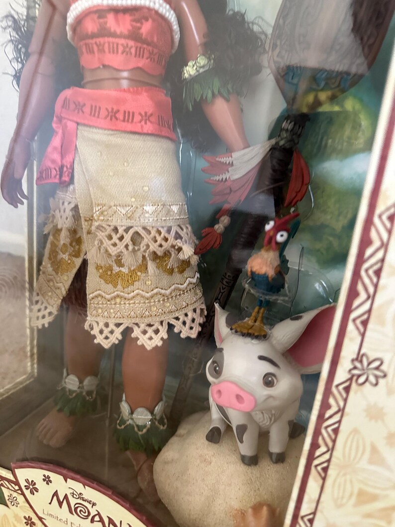 May include: A Disney Moana doll with brown skin, wearing a red and white top, a brown skirt with white trim, and a green lei. She is holding a spear and standing next to a white pig with black spots. The pig is standing on a rock. The background is a brown and white pattern.