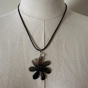 Könnte beinhalten: Eine Halskette mit einem schwarzen Blumenanhänger. Der Anhänger ist eine dunkle, glänzende Blume mit mehreren Blütenblättern. Die Halskette hat eine schwarze Kordel und einen silberfarbenen Verschluss. Die Halskette wird auf einer Schaufensterpuppe präsentiert.