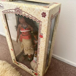 May include: A cardboard box with a clear plastic window showing a Moana doll with brown hair, a red and orange top, a white skirt, and a brown dog. The box has a floral pattern and the text "6+" on the front.
