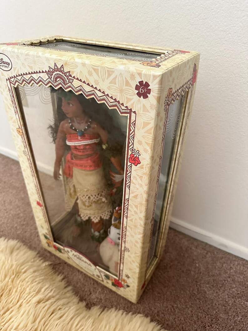 May include: A cardboard box with a clear plastic window showing a Moana doll with brown hair, a red and orange top, a white skirt, and a brown dog. The box has a floral pattern and the text "6+" on the front.