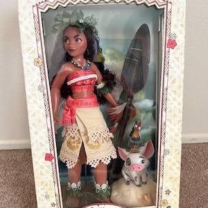 May include: A limited edition Moana doll with a brown and white pig figurine. The doll is wearing a red and white top, a brown skirt, and a flower crown. The doll is holding a wooden paddle with a blue and green design. The box is decorated with a floral pattern and the text "Disney Store" and "Moana Limited Edition 1 of 6500".