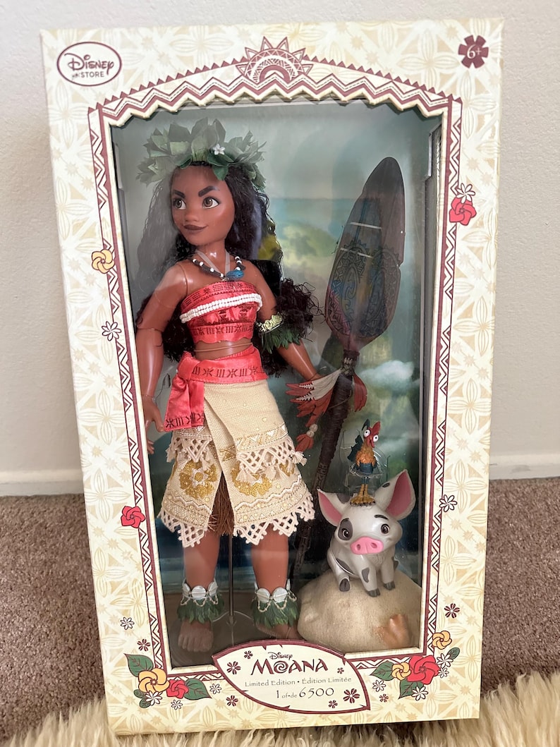 May include: A limited edition Moana doll with a brown and white pig figurine. The doll is wearing a red and white top, a brown skirt, and a flower crown. The doll is holding a wooden paddle with a blue and green design. The box is decorated with a floral pattern and the text "Disney Store" and "Moana Limited Edition 1 of 6500".