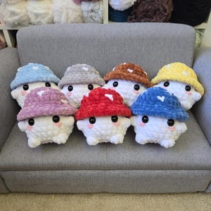May include: A collection of seven plush mushroom figures with white bodies and colorful hats. The hats are in various colors, including blue, red, brown, and yellow, each adorned with white heart-shaped accents. The figures are arranged on a gray sofa.