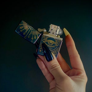 Puede incluir: Un encendedor negro y dorado con un diseño de ojo y un patrón de estallido de estrellas. El encendedor está abierto y la tapa está volteada hacia arriba.