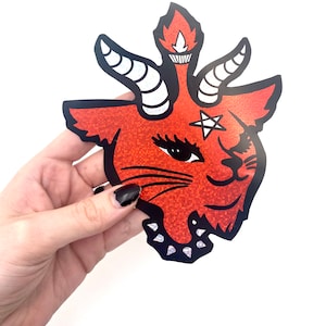 May include: Red glitter cat with a pentagram on its forehead and a flame on its head. The cat is wearing a black choker with silver studs.
