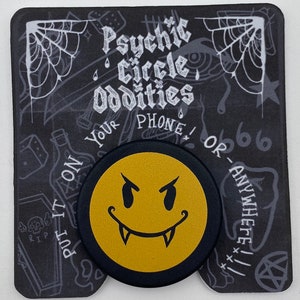 May include: A black and gold phone grip with a yellow smiley face with fangs and angry eyes. The text "Psychic Circle Oddities" is on the background.
