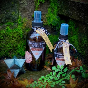 May include: Two brown glass bottles with spray tops and labels that read "Psychic Circle Oddities" and "Grounding Room Spray". The bottles are sitting on a mossy surface with fall leaves and a green plant.