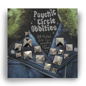 May include: A close-up of a blue denim jacket with 15 silver metal pyramid-shaped pins. The pins are arranged in a random pattern on the jacket. The text "Psychic Circle Oddities 15 Metal Pins for Adorning your Things" is written in black marker on the jacket.